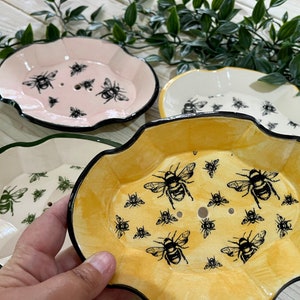 May include: Three ceramic soap dishes featuring bee designs. The dishes are in pink, white and yellow with black bee silhouettes. The dishes have scalloped edges.