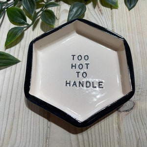 Puede incluir: Un plato hexagonal de cer&aacute;mica blanca con un borde negro. El plato tiene el texto "TOO HOT TO HANDLE" escrito en letras negras.