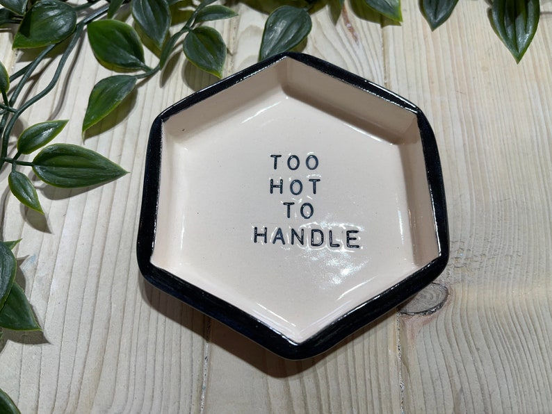 Puede incluir: Un plato hexagonal de cer&aacute;mica blanca con un borde negro. El plato tiene el texto "TOO HOT TO HANDLE" escrito en letras negras.