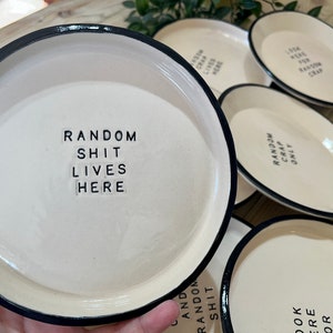Może przedstawiać: Zestaw pięciu białych ceramicznych talerzy z czarnymi obw&oacute;dkami. Każdy talerz ma inny napis wydrukowany czarnymi literami. Napisy to "Random Shit Lives Here", "Random Crap Only", "Look Here For Random Crap", "Random Shit" i "Random Crap Only".