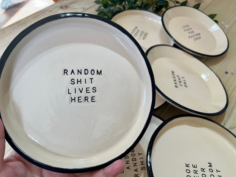 Może przedstawiać: Zestaw pięciu białych ceramicznych talerzy z czarnymi obw&oacute;dkami. Każdy talerz ma inny napis wydrukowany czarnymi literami. Napisy to "Random Shit Lives Here", "Random Crap Only", "Look Here For Random Crap", "Random Shit" i "Random Crap Only".