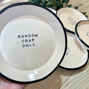 Może przedstawiać: Cztery białe ceramiczne talerze z czarnymi obw&oacute;dkami i humorystycznym tekstem. Talerze mają napis "Random Crap Only", "Look Here For Random Crap", "Lots Crap And Random Crap" i "Some Shit".