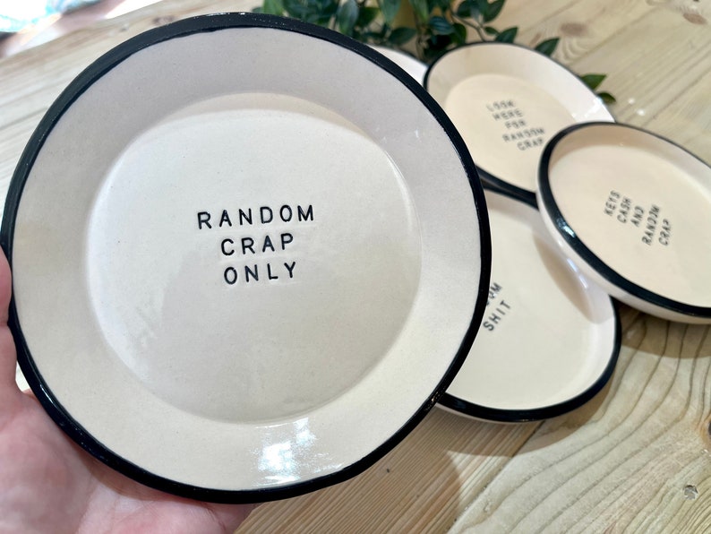 Może przedstawiać: Cztery białe ceramiczne talerze z czarnymi obw&oacute;dkami i humorystycznym tekstem. Talerze mają napis "Random Crap Only", "Look Here For Random Crap", "Lots Crap And Random Crap" i "Some Shit".