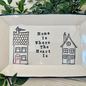 May include: A white ceramic rectangular tray with a black rim. The tray features two black and white illustrations of houses with the text "Home is Where The Heart Is" in black.