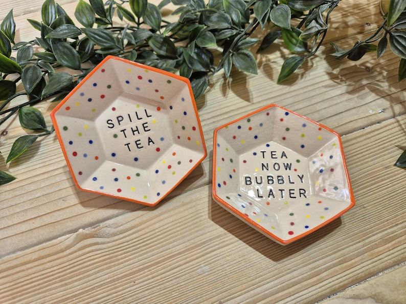 Puede incluir: Dos platos hexagonales de cer&aacute;mica con bordes naranja y fondo blanco moteado. Un plato dice "Spill the Tea" y el otro dice "Tea Now, Bubbly Later".