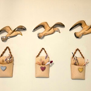 May include: Three wall-mounted ceramic seagull sculptures with three beige fabric wall pockets with rope hangers and dried flowers. Each pocket has a different coloured heart.