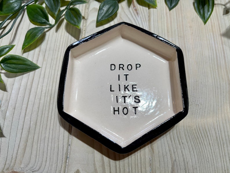 Puede incluir: Un plato hexagonal de cer&aacute;mica blanca con un borde negro. El plato tiene el texto "DROP IT LIKE IT'S HOT" escrito en negro.