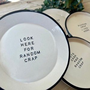 Może przedstawiać: Cztery białe ceramiczne talerze z czarnymi obw&oacute;dkami. Każdy talerz ma inny napis w czarnych literach. Napisy to "Look here for random crap", "Keys and random crap", "Random crap lives here" i "Random crap lives here".