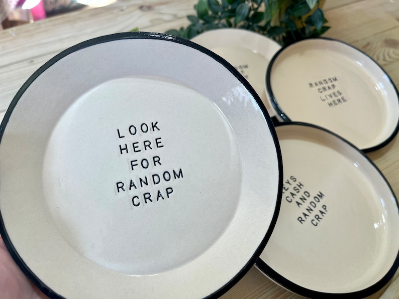 Może przedstawiać: Cztery białe ceramiczne talerze z czarnymi obw&oacute;dkami. Każdy talerz ma inny napis w czarnych literach. Napisy to "Look here for random crap", "Keys and random crap", "Random crap lives here" i "Random crap lives here".
