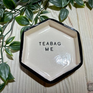 Puede incluir: Un porta bolsitas de t&eacute; de cer&aacute;mica blanca con un borde negro. El porta bolsitas tiene forma de hex&aacute;gono y tiene las palabras "Teabag Me" escritas en &eacute;l.