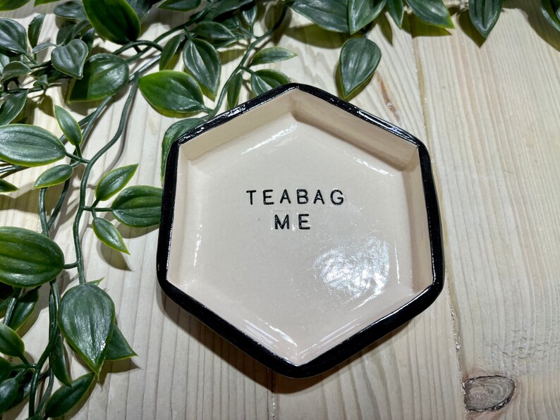 Puede incluir: Un porta bolsitas de t&eacute; de cer&aacute;mica blanca con un borde negro. El porta bolsitas tiene forma de hex&aacute;gono y tiene las palabras "Teabag Me" escritas en &eacute;l.