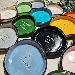 May include: A collection of ceramic dishes with various sayings, including "Stirring Up Trouble", "Stirred Not Shaken", "Stir It Up", and "I'm A Stirrer". The dishes are in various colours, including green, blue, black, grey, and pink.