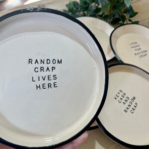 Może przedstawiać: Cztery białe ceramiczne talerze z czarnymi obw&oacute;dkami. Talerze mają r&oacute;żne napisy, w tym "Random Crap Lives Here", "Look Here For Random Crap" i "Keys Cash And Random Crap".