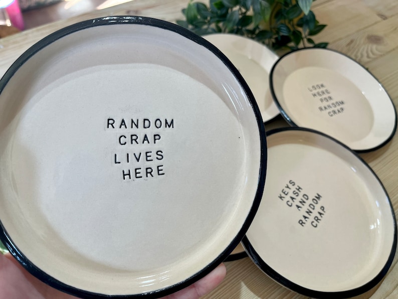 Może przedstawiać: Cztery białe ceramiczne talerze z czarnymi obw&oacute;dkami. Talerze mają r&oacute;żne napisy, w tym "Random Crap Lives Here", "Look Here For Random Crap" i "Keys Cash And Random Crap".