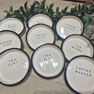May include: A set of cream-coloured ceramic coasters with black rims. Each coaster has black text with phrases such as "Coffee Whore" and "Tea Cunt". Greenery is arranged around the coasters.
