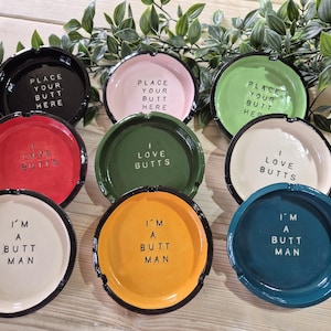 May include: Set of ten ceramic ashtrays with various cheeky phrases printed on them. The ashtrays are in various colours, including green, red, pink, black, white, and yellow.