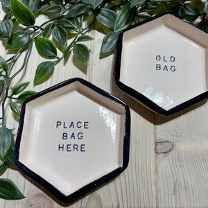 Puede incluir: Dos platos hexagonales de cer&aacute;mica blanca con bordes negros. Un plato dice "Place Bag Here" y el otro dice "Old Bag".