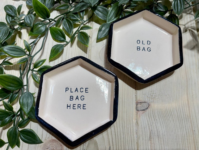Puede incluir: Dos platos hexagonales de cer&aacute;mica blanca con bordes negros. Un plato dice "Place Bag Here" y el otro dice "Old Bag".