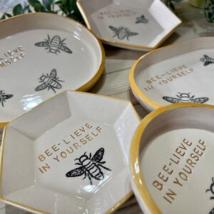 May include: Five ceramic dishes with a gold rim and a white background. Each dish features a bee design and the text "Bee-lieve in yourself".
