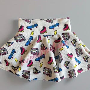 May include: A white skirt with a colorful pattern of roller skates, boomboxes, and skateboards. The skirt has a ruffled hem and is made of a textured fabric.