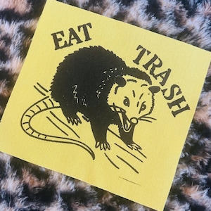 May include: A yellow square patch with the words "EAT TRASH" surrounding a black illustration of an opossum. The opossum is drawn with its mouth open and a long tail. The background is a leopard print pattern.