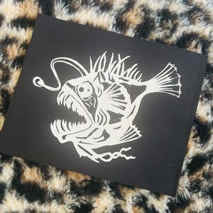May include: A black square patch with a white anglerfish design, showcasing sharp teeth and a glowing lure. The patch is set against a leopard print background, creating a striking contrast.