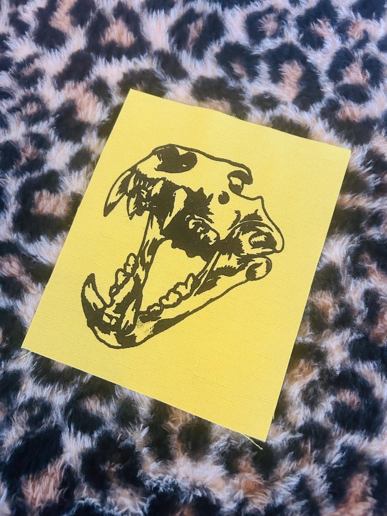 Op de afbeelding: Een gele patch met een zwarte illustratie van een hondenschedel met een open kaak. De patch is rechthoekig en ligt op een achtergrond van luipaardprintstof.