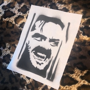 Peut inclure: Pochoir noir et blanc d'un visage d'homme avec une bouche grande ouverte et une expression menaçante. Le pochoir est sur un fond de toile blanche.