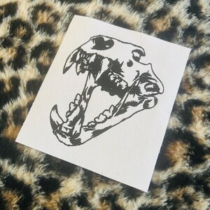 Op de afbeelding: Een witte vierkante patch met een gedetailleerde zwarte illustratie van een hondenschedel met open kaken. De patch is geplaatst tegen een luipaardprint achtergrond. De tanden en botstructuur van de schedel zijn duidelijk gedefinieerd.