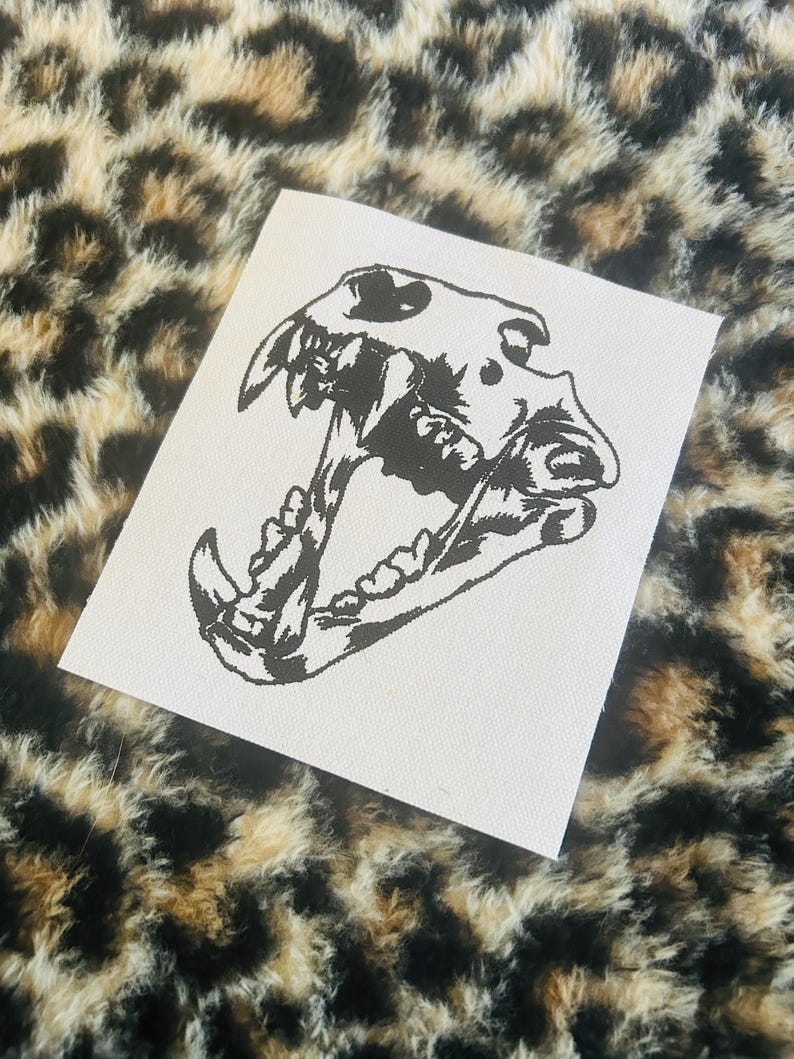 Op de afbeelding: Een witte vierkante patch met een gedetailleerde zwarte illustratie van een hondenschedel met open kaken. De patch is geplaatst tegen een luipaardprint achtergrond. De tanden en botstructuur van de schedel zijn duidelijk gedefinieerd.