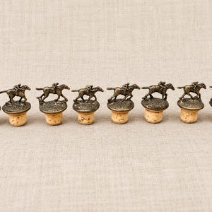 May include: A set of eight bottle stoppers, each featuring a bronze-toned metal figurine of a jockey riding a horse. The metal figures are mounted on circular bases attached to natural cork stoppers, arranged in a horizontal line.