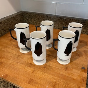 May include: Six white ceramic mugs with black silhouettes of a person wearing a hat and a long coat. The mugs have black rims and handles.