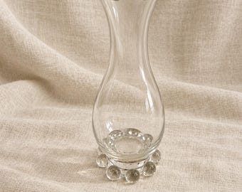 Könnte beinhalten: Eine Vase aus klarem Glas mit einem schlanken Hals und einem gewellten, gerüschten Rand. Der Boden der Vase ist kreisförmig und mit einem dekorativen Ring aus kleinen, klaren Glaskugeln versehen, die am unteren Rand befestigt sind.