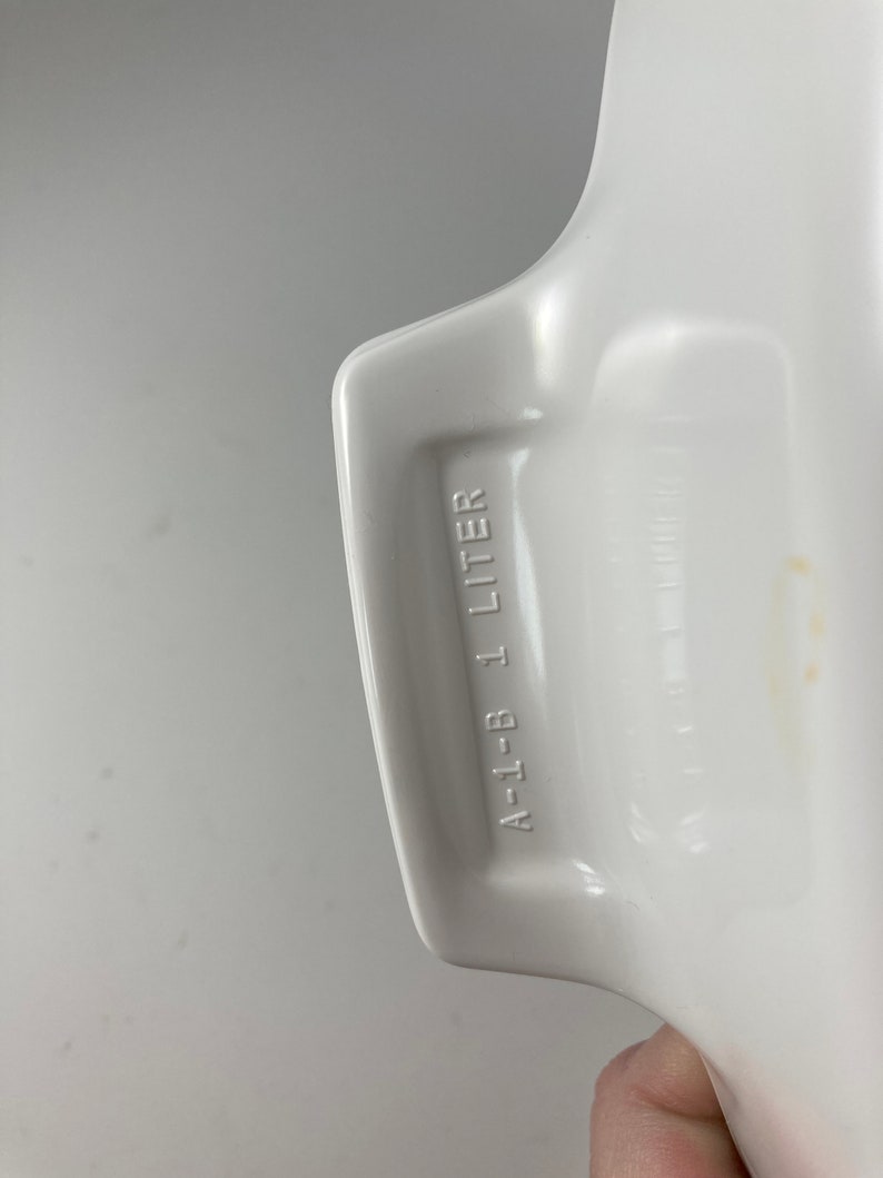 Pu&ograve; includere: Piatto da forno in ceramica bianca con un manico. Il manico ha la scritta "A-1-B 1 LITRO" stampata su di esso.