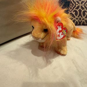May include: A plush lion toy with a vibrant orange and yellow mane and tail. The lion has a tan body and a red heart-shaped tag with the number 2000. The toy is sitting on a light-colored surface.