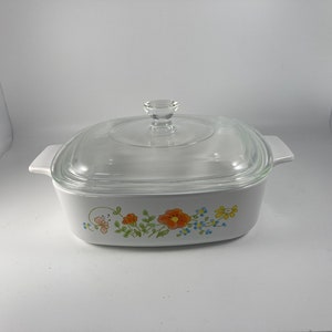 Pu&ograve; includere: Piatto da forno quadrato bianco con coperchio in vetro. Il piatto presenta un motivo floreale con fiori arancioni, gialli, blu e verdi.