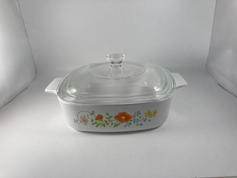 Pu&ograve; includere: Piatto da forno quadrato bianco con coperchio in vetro. Il piatto presenta un motivo floreale con fiori arancioni, gialli, blu e verdi.