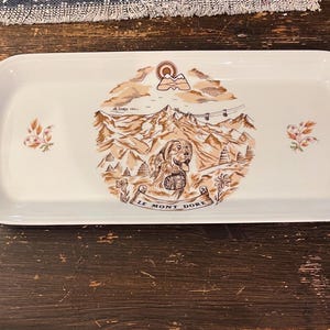 May include: A white ceramic serving tray with a brown and white illustration of a mountain scene with a dog and the text "Le Mont Dore".