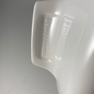 Pu&ograve; includere: Primo piano del fondo di una pirofila in ceramica bianca con le parole "CORNING WARE" stampate sul fondo.
