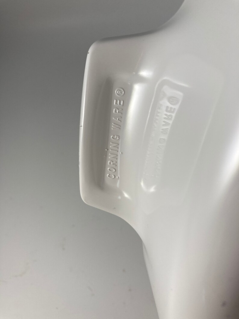 Pu&ograve; includere: Primo piano del fondo di una pirofila in ceramica bianca con le parole "CORNING WARE" stampate sul fondo.