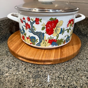 May include: A white enamel cooking pot with a floral design featuring red, yellow, and blue flowers and branches. The pot has a stainless steel lid and sits on a round wooden cutting board. The pot has two white handles.