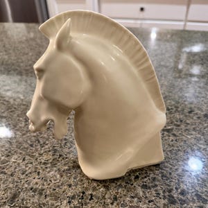 May include: A cream-colored ceramic horse head sculpture. The horse head is sculpted in a classical style, with detailed features including the mane, ear, and facial structure. The sculpture is a solid, three-dimensional form.
