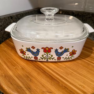 May include: White casserole dish with a clear glass lid. The dish features a decorative band with blue birds, red tulips, and orange flowers. The dish is sitting on a round wooden cutting board.
