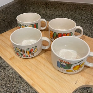 May include: Four white ceramic soup mugs with colorful designs. The mugs have handles and say "Soup" on them.
