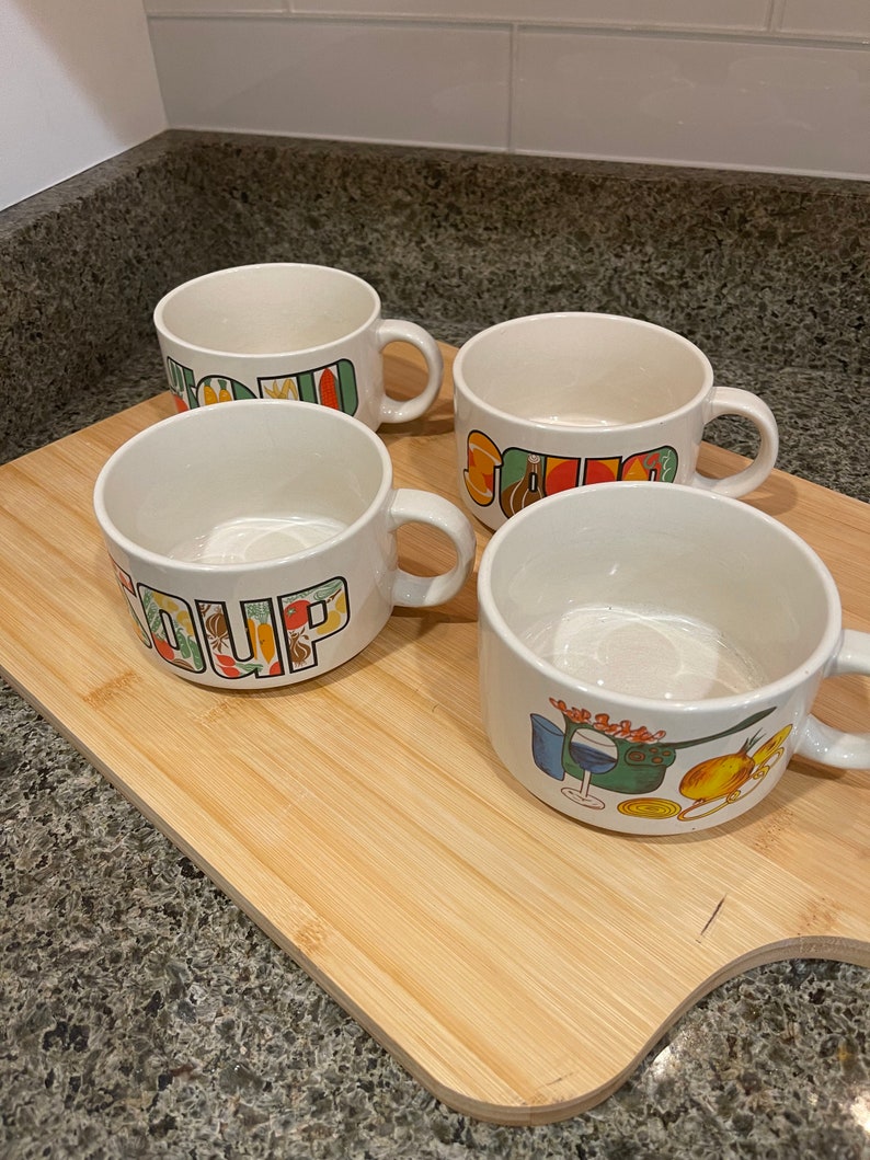 May include: Four white ceramic soup mugs with colorful designs. The mugs have handles and say "Soup" on them.