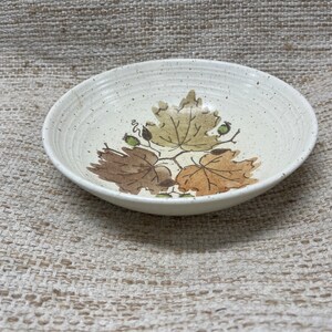 May include: A white ceramic bowl with a speckled glaze features a brown, green, and yellow leaf design. The bowl is perfect for serving snacks or salads.