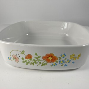 Pu&ograve; includere: Piatto da forno in ceramica bianca con un design floreale con fiori arancioni, gialli e blu. Il piatto ha una forma quadrata con angoli arrotondati e due manici.