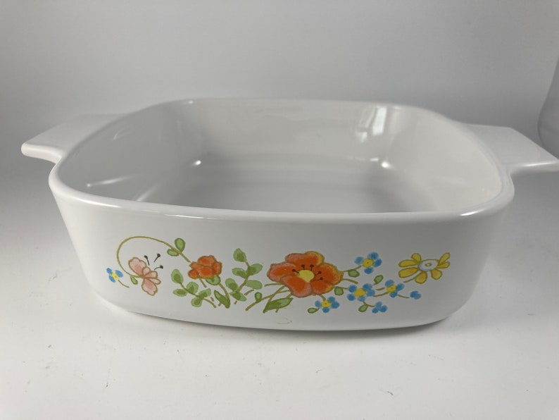 Pu&ograve; includere: Piatto da forno in ceramica bianca con un design floreale con fiori arancioni, gialli e blu. Il piatto ha una forma quadrata con angoli arrotondati e due manici.