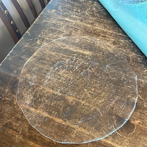 May include: A clear, circular glass plate with a slightly irregular edge sits on a distressed wooden table. A folded, light blue patterned napkin rests in the upper right corner. The table has a dark, aged appearance.