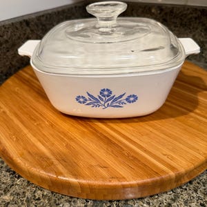 May include: A white casserole dish with a clear glass lid. The dish features a blue floral design and is set on a round wooden cutting board. The background is a dark countertop.